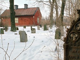 Snötäckt begravningsplats med gravsten i gjutjärn i förgrunden. I bakgrunden står ett rött trähus