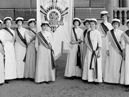 Gruppfoto med kvinnor klädda i vita, fotsida klänningar, studentmössor och ordensband, hållande ett standar