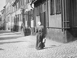 En kvinna med kjol, rutig blus och sjalett går nerför en stenlagd gata med byggnader på båda sidor. Barn och vuxna står eller går också.