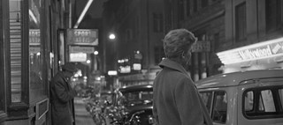 En kvinna och en man står på en upplyst gata i kvällen med ryggen halvt mot kameran