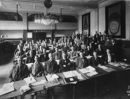 Kostymklädda män sitter i sju rader vid bord med skriv- och ritredskap framför sig. Ytterligare en grupp män står bredvid. De flesta ser in i kameran.