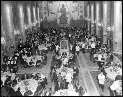 Gyllene salen är full av runda bord med sittande gäster. Vid sidan finns en orkester och en kör. Servitörer rör sig mellan borden.