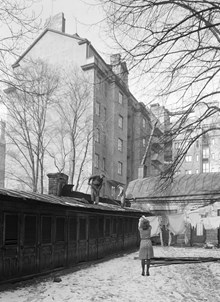 Kammakargatan 70, gården. En kvinna hänger tvätt och två män arbetar på taket till längan med utedass. Huset i fonden ligger vid Tegnérgatan 57
