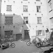 Fasaden till Sven Vintappares Gränd 2A med budcyklar, cyklar och mopeder parkerade på den öppna platsen framför byggnaden.