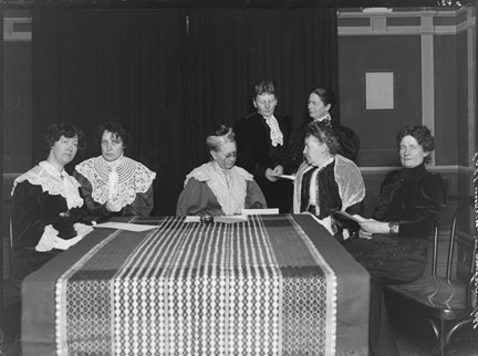 Fem kvinnor sitter vid ett bord och två står bakom. Flera bär stora spetskragar.
