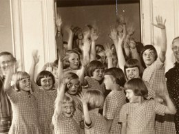 En grupp kvinnor och barn i rutiga klänningar står i en dörröppning och vinkar