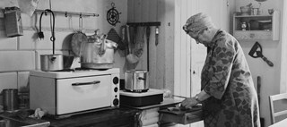 En elektrisk spis med stor kaffekittel på i ett äldre, omodernt kök. En kvinna skär upp någonting