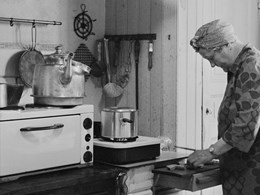 En elektrisk spis med stor kaffekittel på i ett äldre, omodernt kök. En kvinna skär upp någonting