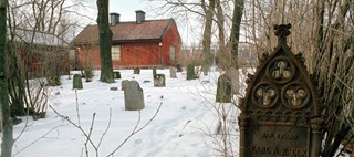 Snötäckt begravningsplats med gravsten i gjutjärn i förgrunden. I bakgrunden står ett rött trähus