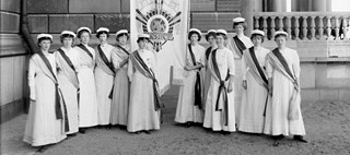 Gruppfoto med kvinnor klädda i vita, fotsida klänningar, studentmössor och ordensband, hållande ett standar
