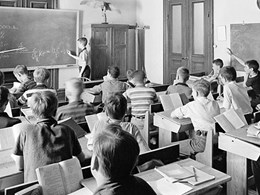 Klassrum med elever i bänkarna och en lärare med pekpinne.
