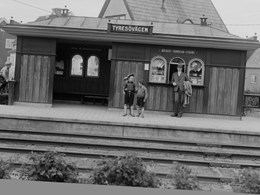Tre barn och en herre i hatt står vid en enkel träbyggnad i ett villakvarter
