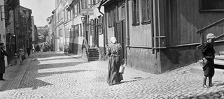 En kvinna med kjol, rutig blus och sjalett går nerför en stenlagd gata med byggnader på båda sidor. Barn och vuxna står eller går också.