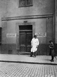 En man och en flicka står vid porten till Bellmansgatan 15. 2 tr. upp finns skomakare J.W. Larsson. Dåvarande kv. Sankta Maria, nu kv. Magdalena