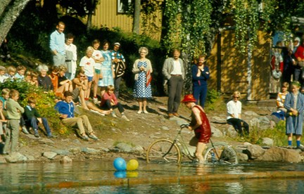 Färgfotografi på barn och personal på klippa samt cykel i vattnet på Barnens Ö.