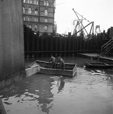 Två män i en farkost på Riddarholmskanalen. Byggkranar i bakgrunden.