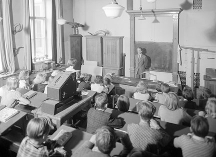 Svartvitt foto som visar ett klassrum med elever som sitter i sina bänkar och en manlig lärare som står vid svartatavlan