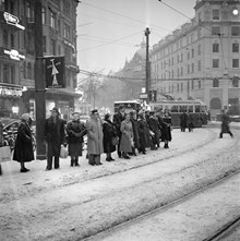 Stureplan och Birger Jarlsgatan söderut. Stureplan 2 och 3. Folk väntar på buss/spårvagn i snöyran. På två stolpar sitter affischer med ett snapsglas och ett ljus, ""Släck inte julglädjen""
