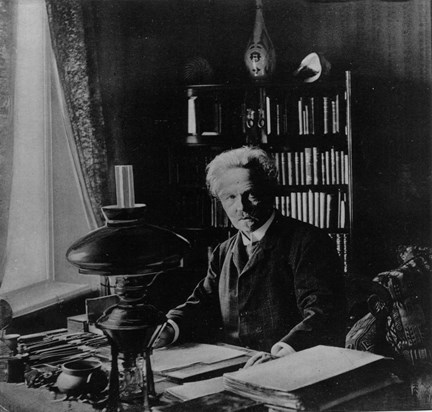 August Strindberg sitter vid sitt skrivbord. På bordet högar med papper, skrivdon, en fotogenlampa med mera. I bakgrunden syns en bokhylla med böcker, en hög keramikvas och två stora snäckor