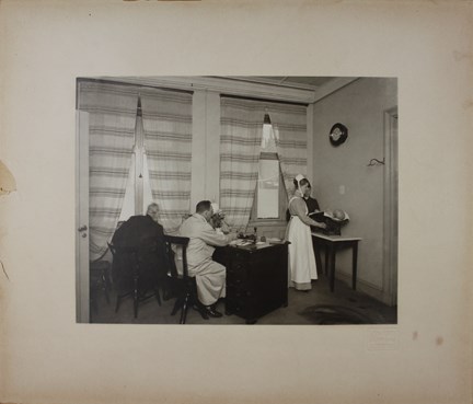 I ett rum med randiga gardiner för fönstren sitter två män vid ett skrivbord medan en sjuksköterska väger ett litet barn och en kvinna tittar på.