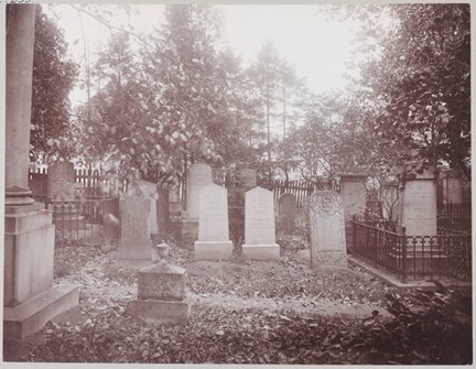Ett svart-vitt fotografi på en begravningsplats med minst 13 synliga gravstenar. Staket omger några av gravstenarna och ett högre staket i bakgrunden omger hela platsen. Höga träd syns i bakgrunden.