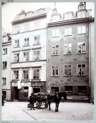 Två äldre stenhus med ett torg belagt med gatsten framför. På torget står en häst med vagn. Intill vagnen står en man med en väska i handen.
