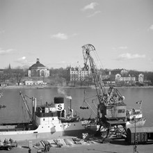 Kranen lyfter tunnor till en ångbåt vid Skeppsbrokajen. Skeppsholmen i fonden