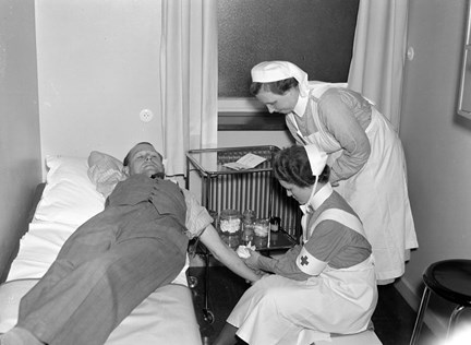 En man ligger på en brits. Två sjuksköterskor och en vagn med material för blodgivning intill.