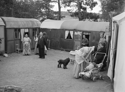 I bilden syns åtta vuxna människor, två barn och en hund. Runtomkring syns tält-bostäder och bakom tälten träd.