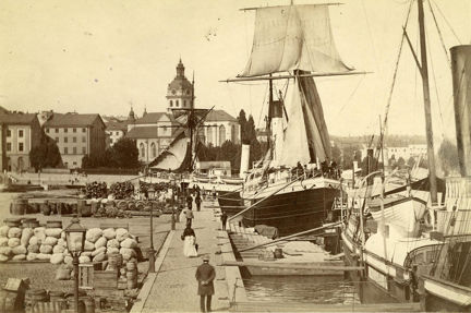 Fotografi på Skeppsbrokajen 1873. Vid kajen ligger fartyg samt säckar och tunnor med varor. 