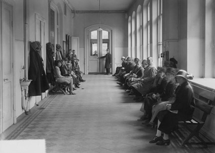 I en korridor med mönsterlagt golv och ljusinsläpp från höga fönster sitter väntande människor på rad längs väggarna.
