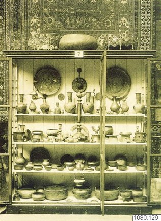 Svartvitt fotografi som visar en monter med metallkärl och keramik. I bakgrunden en persisk matta.