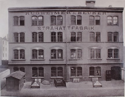 Svartvitt fotografi på fasad med texten Rügheimer & Becker stråhattsfabrik.