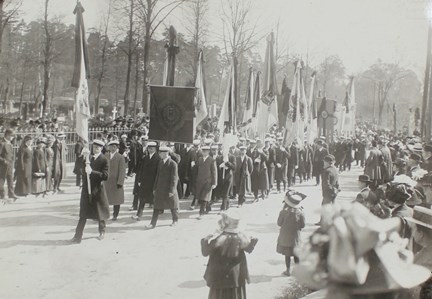 Långt begravningståg med flaggor och standar samt åskådare vid sidan om.