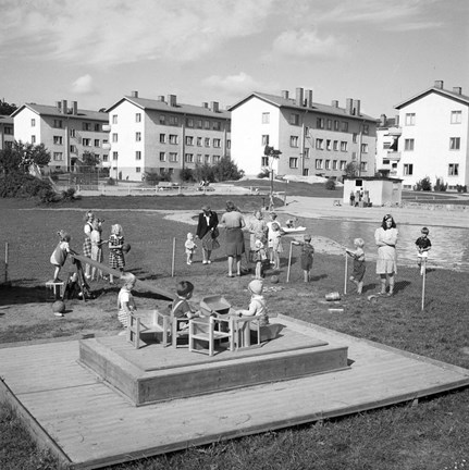 Framför fyra flerfamiljshus i tre våningar ser vi en äng med en stor lekplats. Till höger finns en större plaskdamm, till vänster leker barn på ett trädäck med små stolar och bord. I bakgrunden syns en gungbräda.