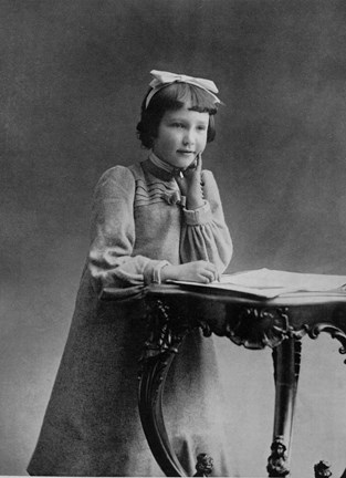 Ateljéfotografi av Kerstin Strindberg i 8-årsåldern, stående vid en pulpet, klädd i ljus klänning med puffärmar och veck och med en rosett i håret.