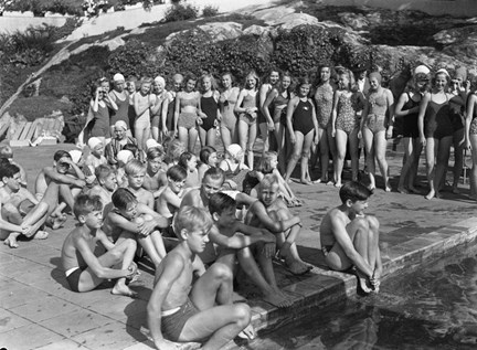Intill kanten av en simbassäng sitter en grupp om ett tjugotal pojkar på marken. Intill står ungefär lika många flickor. De är mellan uppskattningsvis 8 och 13 år.