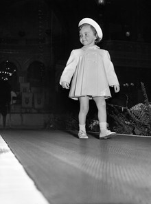 En flicka i kappa och hatt på catwalken under Röda korsets barnmannekänguppvisning