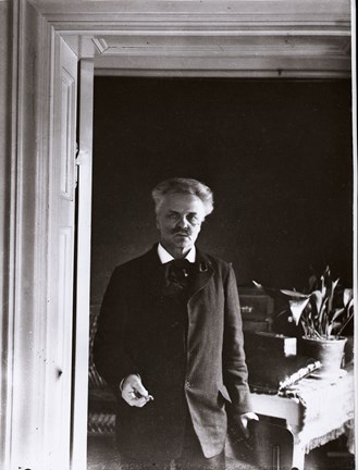 August Strindberg står i en dörröppning, iklädd mörk kostym och kravatt och med en cigarett i handen. I bakgrunden ett bord med duk, en lerkruka med en växt.