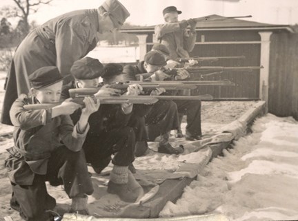 Unga pojkar tränar knästående skytte under ledning av en militär.