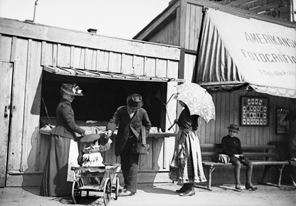Marknadsstånd på Skansen med marknadsbesökare och barn i skrinda. 
Till höger under markisen har "Amerikanska - Fotografi Ateliern" sin fotoateljé.