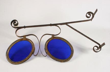 Skylt som suttit fast i fasaden, i form av glasögon med blått glas.