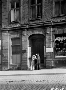 Två kvinnor står utanför porten till Hornsgatan 1. I skyltfönstret hänger löskragar och skjortbröst. Ovanför porten sitter en skvallerspegel i fönstret