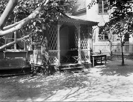Ett lusthus med en veranda, ett träd växer precis intill. Vid sidan står ett bord, skuggor av träd på marken.