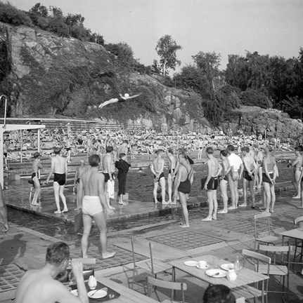 En kvinna gör ett elegant simhopp ned i en simbassäng. Människor i och runt poolkanten tittar på. I bakgrunden syns en brant bergvägg.