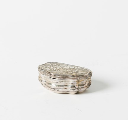 Stängd snusdosa i silver fotograferad snett från sidan, locket har böljande kanter och en mönstrad yta.
