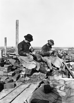 Svart-vit bild med två män som slår tegelstenar med hammare i händerna. Männen bär hatt, skjorta, byxor, förkläde och rejäla skor. I bakgrunden utsikt från hög höjd över staden. Runtom männen ligger tegelstenar.