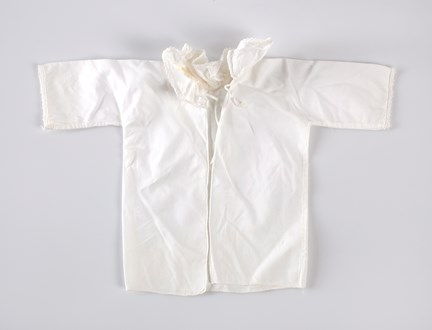 Babyskjorta av vitt bomullstyg, knyts med två band baktill. Uddspets längs kragens kant och längst ner på ärmarna.