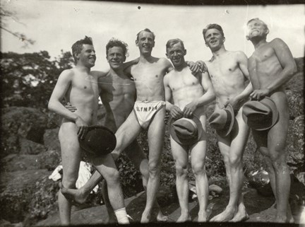 Foto på sex unga män  på klippa. En av dem är iförd Olympias badbyxor. De övriga är nakna men skyler sig med hattar som de håller framför sig. 