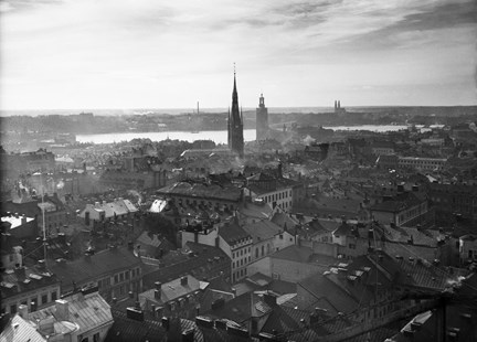 Utsikt från ett högt hus, södra Kungstornet. En stor mängd hustak syns, liksom flera kyrktorn. I fonden ligger vattnet Riddarfjärden och Stockholms stadshus med sitt höga torn. 